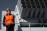 A man in a navy uniform and life jacket stands beside a rocket launcher on a ship.