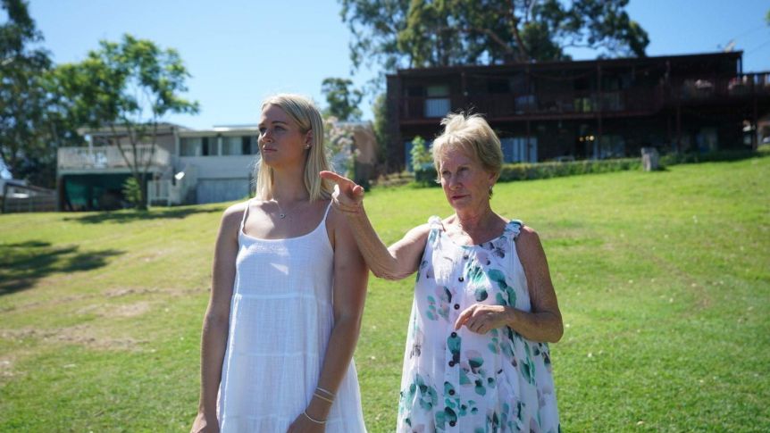 Two women in white dresses stand on a grassy hill.